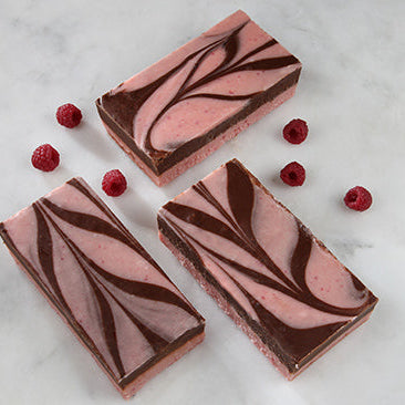 Three pieces of raspberry chocolate swirl fudge with chocolate on top and raspberries on the side, placed on a marble surface.
